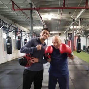 Two men in boxing gloves in a gym, one young and one elderly, smiling after training.