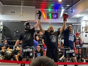 Muay Thai fighters celebrating victory in gym with raised arms and gloves.