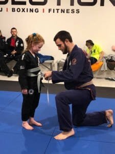 Young girl receiving belt promotion from instructor in Brazilian Jiu-Jitsu class.