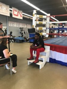 Young boxer being interviewed in boxing gym with ring and equipment.