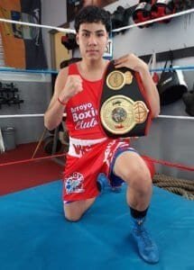 Young boxer in red gear holding championship belt in boxing gym.
