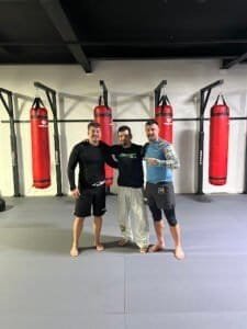 Three men in boxing gym with punching bags, smiling and posing for photo.