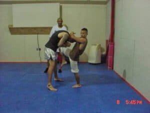 Two fighters practicing MMA techniques in a training gym with a coach observing.