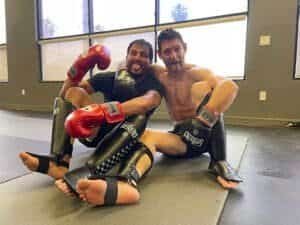 Two Muay Thai fighters sitting on the mat with boxing gloves and shin guards.