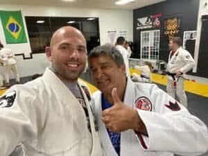 Two men in Brazilian Jiu Jitsu gis smiling at the camera in dojo.