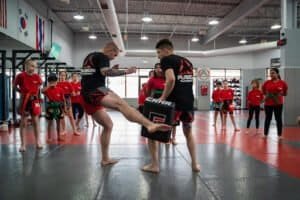 Martial arts class with instructor demonstrating kick to student in gym.