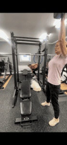 Woman lifting weights with trainer and dog in gym.