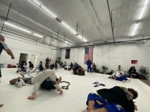 Jiu Jitsu practitioners training in a spacious gym with white mats and American flag.