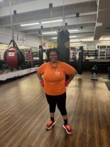 Woman in orange shirt and boxing gloves in boxing gym with punching bags.