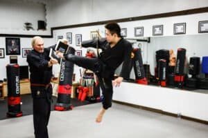 Martial arts student practicing high kick with instructor in dojo.