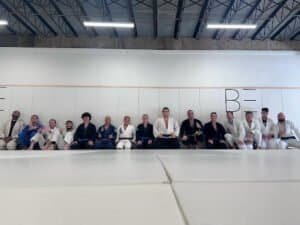 Group of Jiu Jitsu practitioners sitting on mats in dojo.