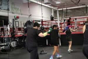 Women practicing boxing techniques in a gym with a boxing ring and training equipment.