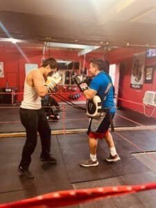 Two men sparring in boxing gym with gloves and punching bags.