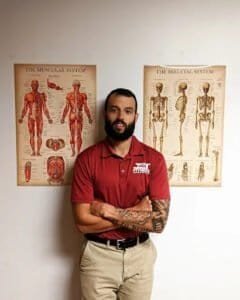 Fitness trainer standing with arms crossed in front of muscular and skeletal system posters.